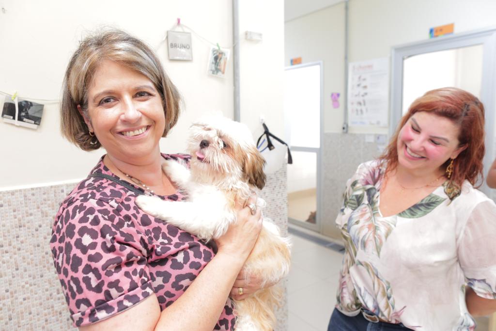 Na imagem a Sandra Sabino está segurando um cachorro e sorrindo para a foto. Ela tem cabelos curtos e usa camisa rosa com detalhes pretos. Ao seu lado está a Analy Xavier olhando para o cachorro e sorrindo. Ela usa uma camisa branca com detalhes verdes.